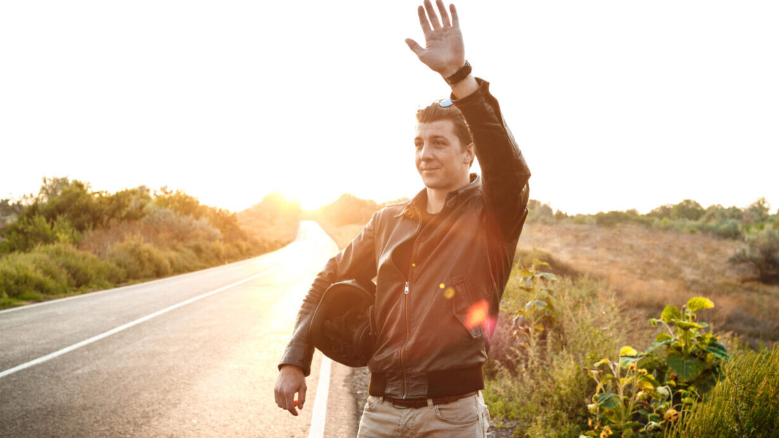 Young handsome motorbiker in leather jacket greeting, holding helmet, posing at countryside road.