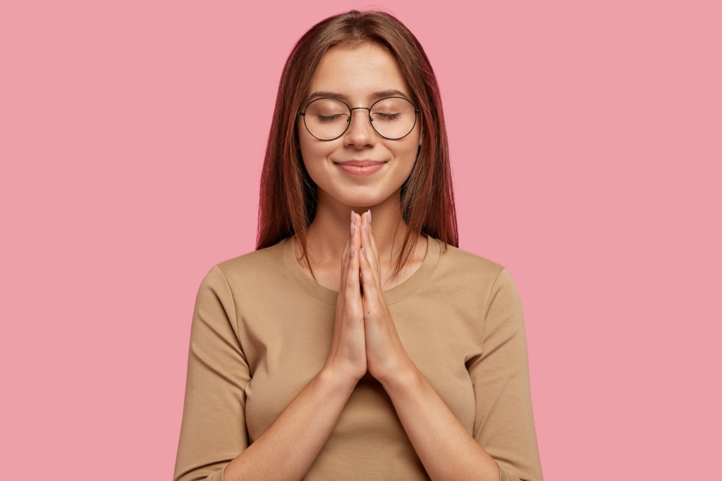 Beautiful European woman beggs for help or asks for apologize while keeps hands in praying gesture, wears round spectacles, has dark hair, dressed in casual outfit, isolated over pink background