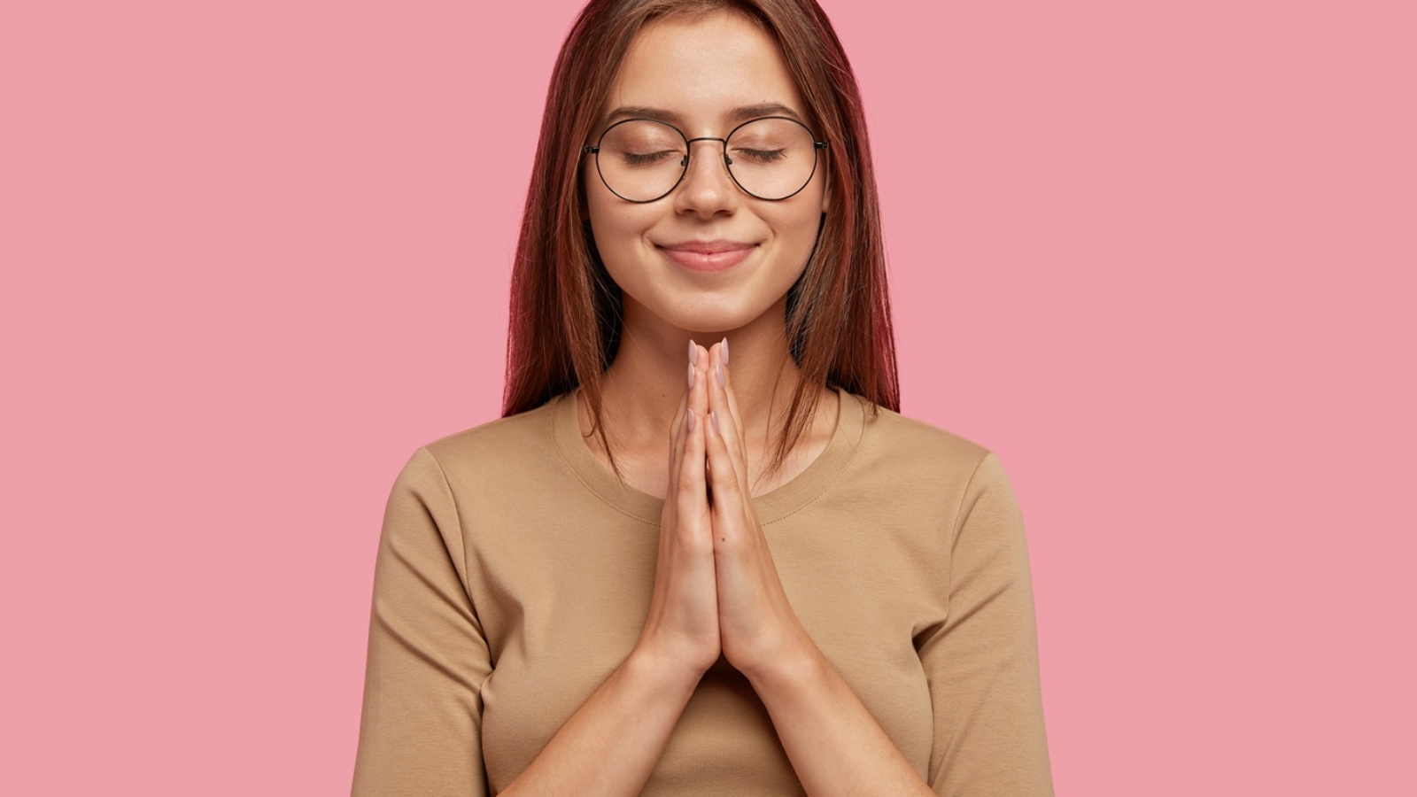 Beautiful European woman beggs for help or asks for apologize while keeps hands in praying gesture, wears round spectacles, has dark hair, dressed in casual outfit, isolated over pink background