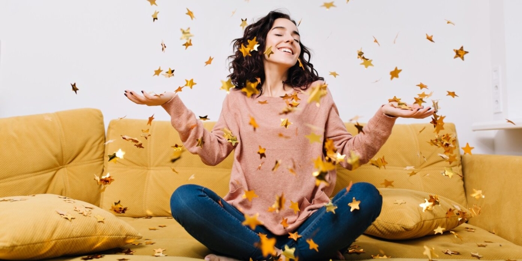 Expressing true positive emotions of young joyful woman with cut curly hair having fun in falling tinsels on couch in modern apartment. Home cosines, joy, smiling