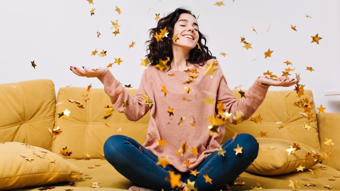 Expressing true positive emotions of young joyful woman with cut curly hair having fun in falling tinsels on couch in modern apartment. Home cosines, joy, smiling