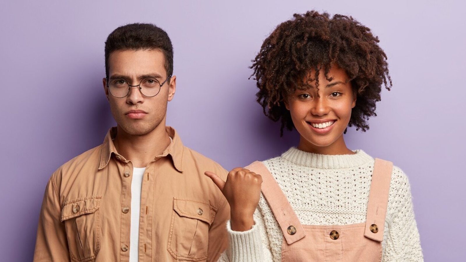 Young diverse couple pose together against purple background. Positive beautiful Afro American woman wears white sweater and overalls, points with thumb at serious dissatisfied guy in spectacles