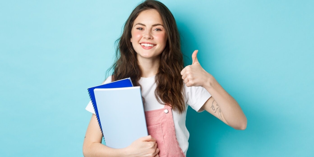Young woman attend courses, girl student studying, holding notebooks and showing thumb up in approval, recommending company, standing over blue background.