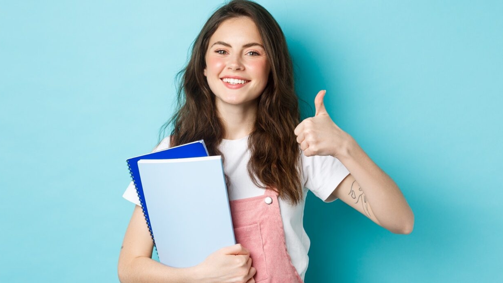 Young woman attend courses, girl student studying, holding notebooks and showing thumb up in approval, recommending company, standing over blue background.